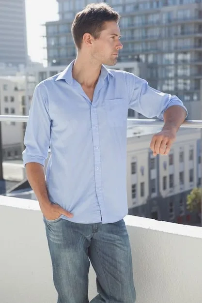 A young man wearing a light blue button-down shirt over a white t-shirt, paired with dark wash jeans and white sneakers, suitable for a casual spring day. He is carrying a lightweight jacket over his arm.