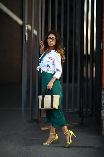 A woman in a stylish, deep green pantsuit, switching from sensible office heels to strappy, embellished high heels, with a small clutch bag replacing her larger work tote.