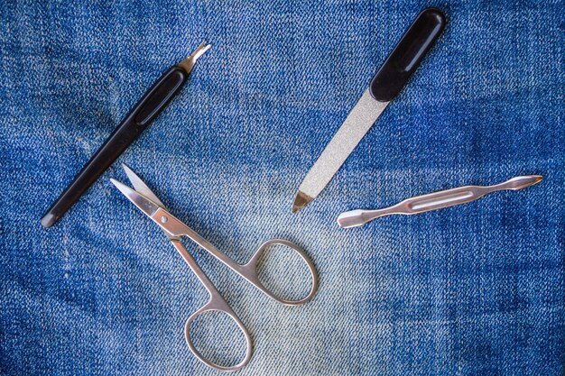 A flat lay of various distressing tools including sharp scissors, a utility knife, sandpaper, a cheese grater, and tweezers, alongside a pair of blue denim jeans.