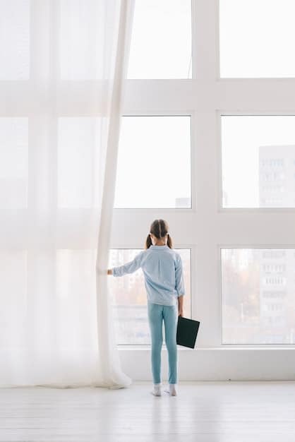 A bright living room with floor-to-ceiling sheer white curtains pulled back to reveal large windows, light wooden floors, and a minimalist design.