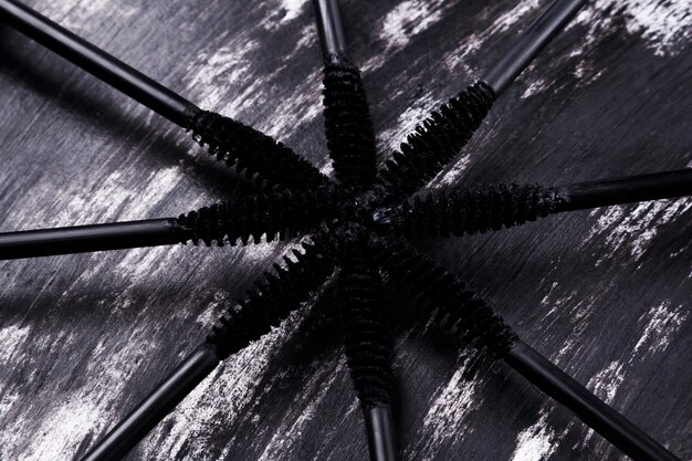 A detailed close-up shot of an open tube of mascara with the brush pulled out, showing the bristles coated with black product. The background is a soft, out-of-focus array of other cosmetics.