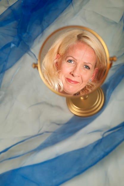 A woman in her 60s looking thoughtfully at her reflection in a mirror, her skin appears well-cared for, conveying self-love and confidence.