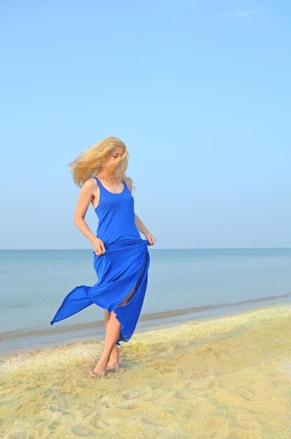 A woman wearing a flowing linen maxi dress in a vibrant blue color, walking barefoot on a sandy beach with the ocean in the background. Her hair is loosely tied, and she wears simple, natural jewelry.