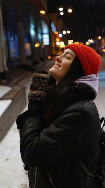 A person in a chic oversized black wool coat, wearing a thick knit scarf and a beanie, walking on a snowy city street with blurred city lights in the background.