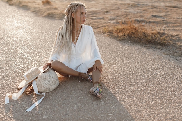 A stylish model wearing light wash wide-leg jeans with an embroidered white peasant blouse, espadrille wedges, and carrying a woven straw bag, exuding a relaxed, bohemian summer vibe.