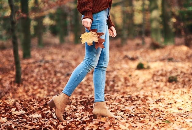 A person walking in an autumnal park, wearing dark-wash straight-leg jeans, a chunky knit cream sweater, a tan trench coat, and brown leather ankle boots, with fallen leaves on the ground.