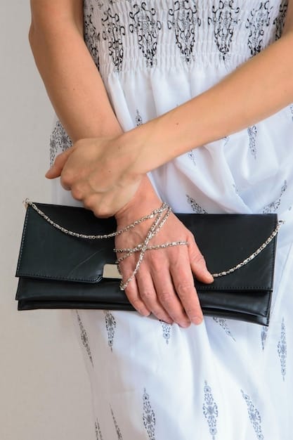 A close-up shot of a stylish woman's hands adjusting a delicate gold layered necklace over a simple white tee, with a sleek, structured handbag resting on her lap.